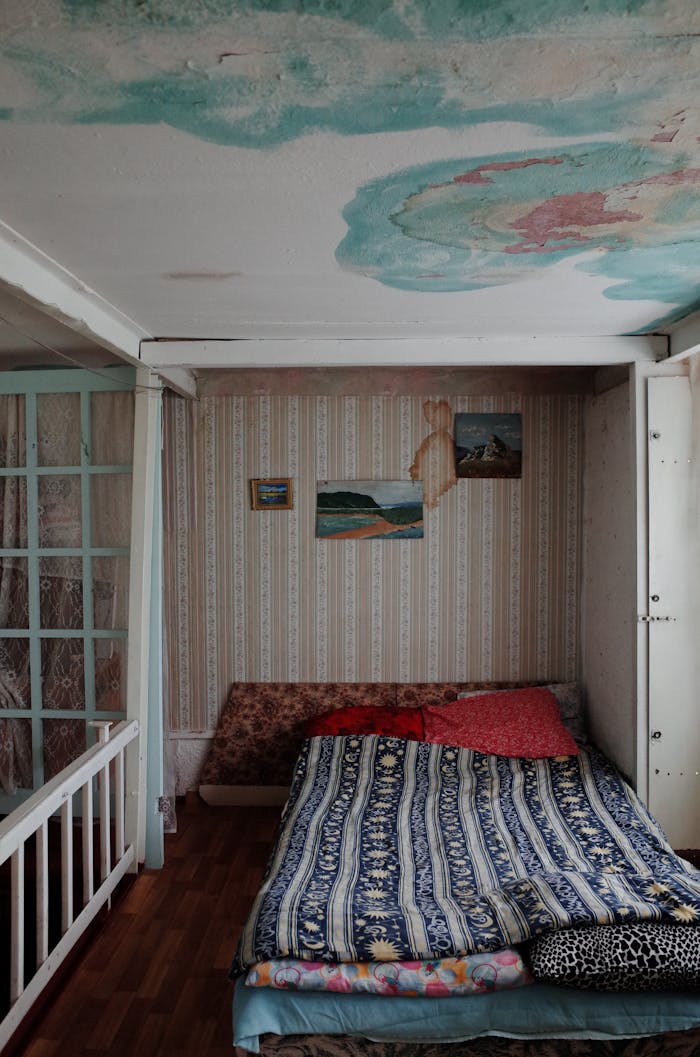 gallery-4 A small bedroom with a unique ceiling design, featuring water stains and an eclectic bed setup.